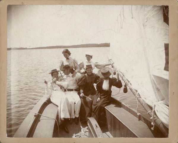 A young man with cap and light-colored tie is sitting in the stern of a sailboat with five young women. He has his arms around two of the women; two others are resting a hand on the boom. A caption written on the reverse of this image identifies "Alfred J. Brumder and his girl friends on the sloop 'Henrietta' about 1895."  