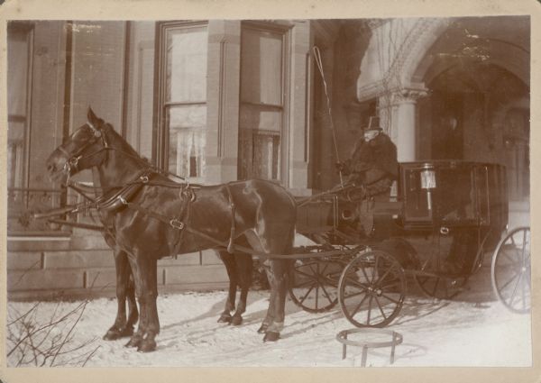 A team of two horses is harnessed to a highly polished carriage with a side lamp. The driver is holding a long whip and is wearing a top hat, winter coat and a fur capelet. The rig is parked in front of a bay window, just beyond the porte-cochere of a large stone house. There is snow on the ground.