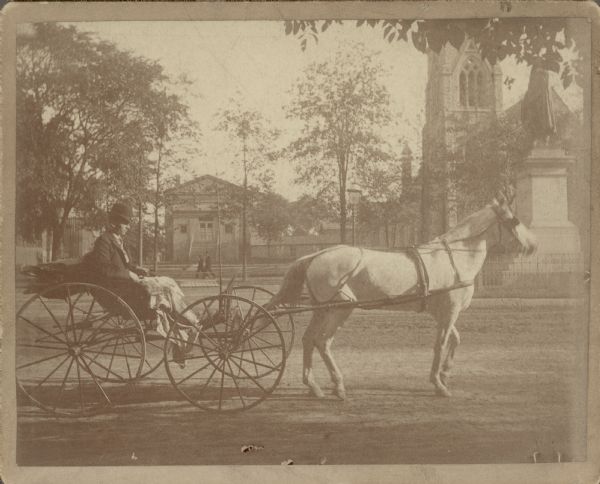 A light-colored horse is hitched to a small buggy driven by Lester Carr. Carr is wearing a hat and has a lap robe across his legs. The buggy is stopped on Grand Avenue (since 1926, Wisconsin Avenue) near the George Washington Monument. St. James Episcopal Church is in the background, right. A caption on the reverse states: "Our horse 'Dolly' with Lester Carr in buggy. This picture is taken by Alfred Brumder, as were all those mounted like this one." 