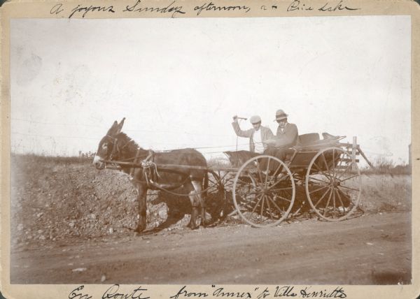 Two men are riding in an open carriage pulled by a donkey. Captions written on the front of the photograph describe "A joyous Sunday afternoon at Pine Lake," and "En Route from 'Annex' to Villa Henrietta." One man, identified only as "Goldsmith," is holding a whip, while the other man, likely Herman Otto Brumder, is holding the reins. The carriage is stopped on the shoulder of a road. On the reverse is written: "I think that donkey had too many "High Balls" that day for he wouldn't move. Don't you wish you'd been there. With much love. H.O.B. [Herman Otto Brumder]" In different handwriting is the note: "The 'Annex' was a small tavern with a bar and two tables only. It was part of the Interlaken Hotel on Beaver Lake."  