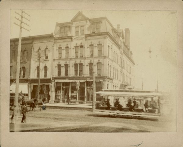 View from street towards a streetcar passing in front of the 1872 Brumder building located, according to a note of the reverse of the photograph, "on N. E. corner of Plankington [sic] Ave. and Kilbourn." When the building was constructed, N. Plankinton was called West Water Street. George Brumder's bookstore and Germania Printing offices were located on the first floor of this building; the family lived on the second floor. There are pedestrians on both sides of the street and a horse-drawn wagon on the left.