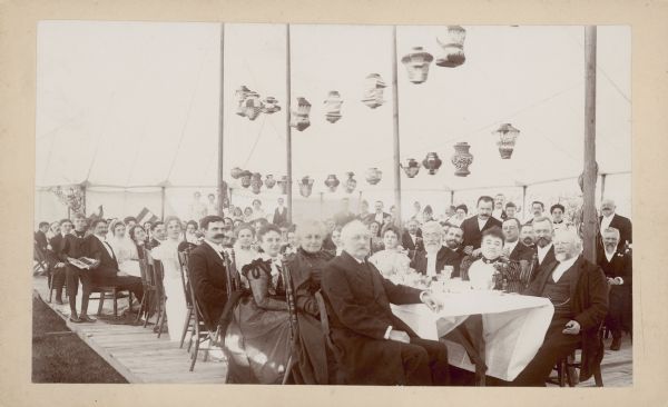 Well-dressed men, women and children are sitting at long tables, and more people are standing in the background for a group portrait at a wedding reception. They are under a large tent with a temporary wooden floor. Paper lanterns are strung overhead. A boy wearing a sailor outfit is standing on the left holding an open box of cigars. He is next to a young man and woman whose chairs are pulled back from the table; the woman is wearing a veil and is most likely the bride. A caption written on the reverse identifies the married couple as Lorle Meinecke and [Dr.] Onno Boekhoff of Milwaukee. A short notice in an 1898 medical journal states that the wedding took place in Wauwatosa.  