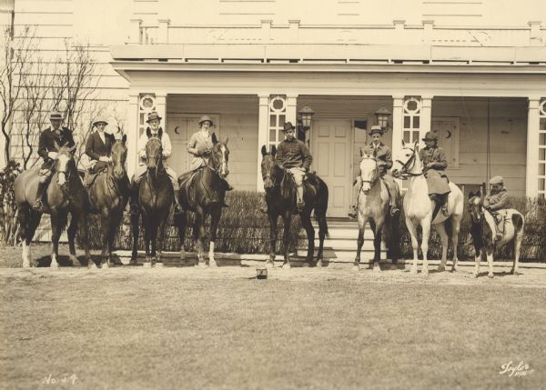 Walter Vail Johnston, far left, and his wife June Crandall Johnston, second from left, posing with their six children in front of their large house on Dean Road. The entire family is on horseback, with the youngest son, John Keith Crandall Johnston, at far right on a pony. The others are, from third left, left to right, "Wallie," Walter Vail Johnston Jr.; "Sis," June Ellen Johnston (Mrs. William George) Brumder; "Ned" Edward Crandall Johnston; David Crandall Johnston; and "Patsy," Patricia Johnston, later Mrs. Harald "Shorty" Pabst. The men and boys are wearing jodhpurs and the women are riding side saddle. The horses have English saddles. A camera case is lying on the ground in front of Ned. The names of several of the horses are also included in a caption on the reverse of the photograph.
