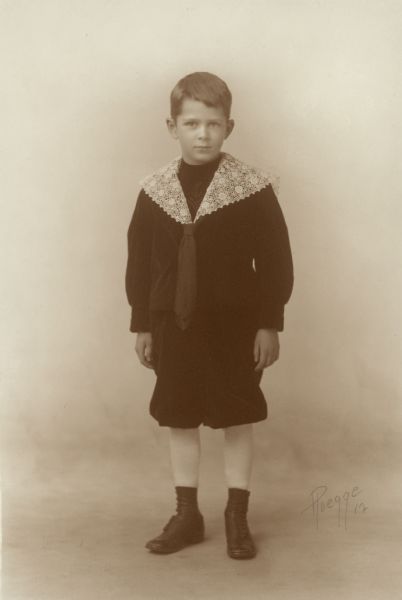 Edward Brumder posing for a full-length, standing studio portrait. He is wearing knickers and a sailor style top with kerchief and fancy lace collar.