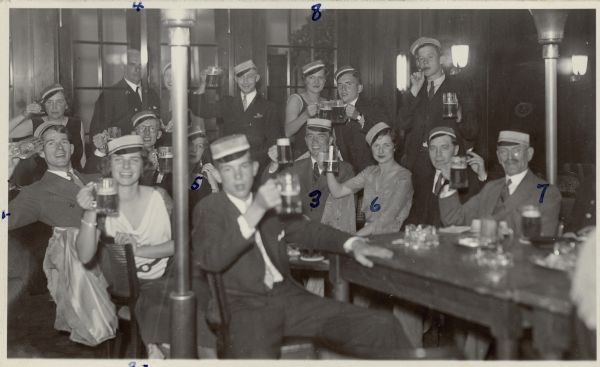 A group of mostly young people are sitting and standing around wooden tables, raising steins of beer while posing for a group portrait. Nearly all of the group are wearing caps with small visor-like brims typical of German (male) student societies. Eight members of the group are identified by numbers written in ink on the front of the postcard. A legend and caption on the reverse identifies, " 1 + 2 = Ed + Marion Brumder; 3= H. de Give; 4 = Capt. Brümings; 5 = Adele Bronhoff; 6 = Dorothy Corbett; 7 = Mr. Gussman; 8 = 'Fritzie'.  Student nite [sic] aboard S.S. <i>Von Steuben</i> - mid-Atlantic - Capt. Brümings in rear..."