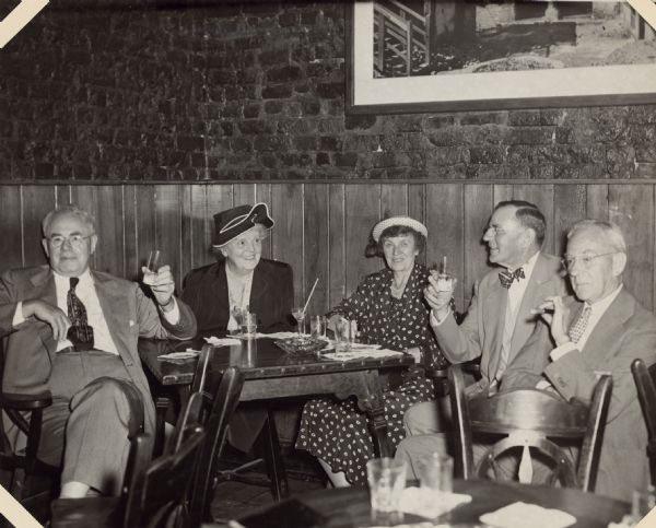 Six well-dressed patrons are posing sitting around a wooden table at Pat O'Brien's Bar in New Orleans. They are identified as, left to right: Walter A. and Toni Zinn; Marnie (Margaret Bouer) and Herbert Paul Brumder; and George F. Brumder. George is holding a cigar in a holder; the other two men are lifting their drinks. There is wood paneling on the lower part of the wall behind them with exposed brick above.
