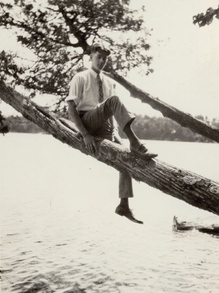 Ed Brumder is sitting on the branch of a tree which has grown out over the water of Pine Lake. The far shore is in the background.