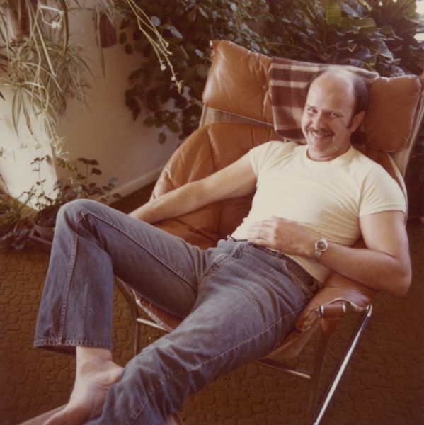Edward John Brumder Jr. is casually reclining in a contemporary style leather and chrome easy chair in his apartment in Juneau Village. He is barefoot, and is wearing bell bottom jeans and a white t-shirt. There are houseplants behind him.