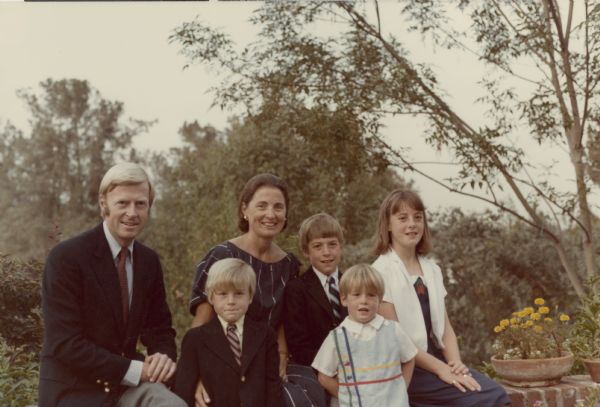 George Albert and Marilyn Ann (Coyne) Brumder are posing sitting outdoors on a brick wall with their four children. From left: James Frederick, George Richard, Stephen Gallun, and Thekla Henrietta. There are trees in the background and a pot of marigolds on the right. George Albert Brumder, an attorney and civic leader in Pasadena, California, is a great-grandson of prominent Milwaukee publisher George Brumder.