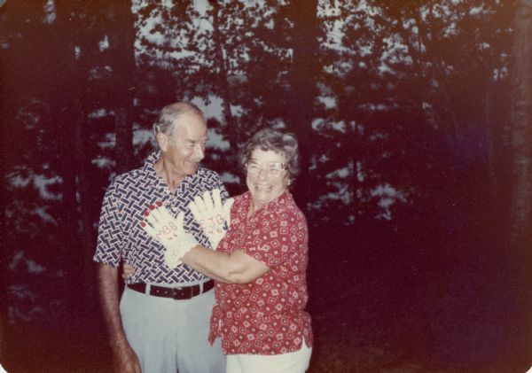 Edward John and Marion (Briggs) Brumder posing for a snapshot on their forty-sixth wedding anniversary. They are casually dressed and each is wearing and showing off a decorated oven glove which features a wedding ring. Marion's glove has lace trim, painted fingernails and her initials. Ed's glove has his initials and an anchor. Pine Lake can be glimpsed through the trees behind them. 