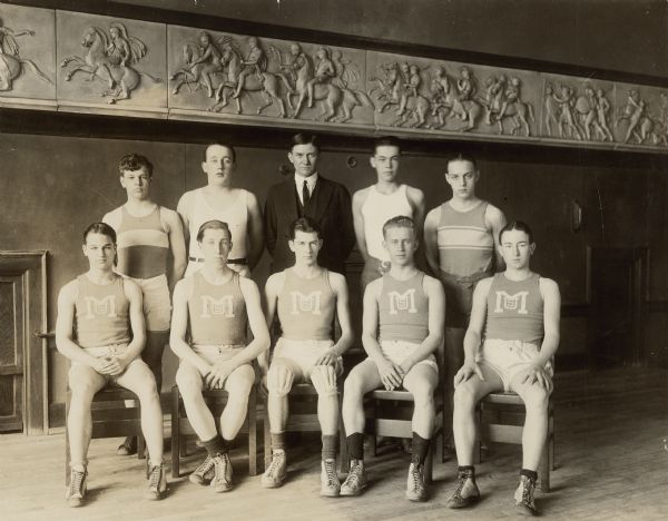 Edward John Brumder, center front with knee pads, posing for a team portrait with his teammates and coach in the gymnasium of the Milwaukee University School. There is a reproduction classical bas-relief frieze on the wall. A caption on the reverse of the photograph also identifies "Joe Gutenkunst top right." Edward was the son of George Frederick Brumder and grandson of George Brumder, founder of the Germania Publishing Company.