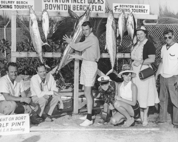 Edward J. Brumder, Jr., center, posing with a sailfish which is hanging on a rack with other fish. A sign identifies the location as the Boynton Inlet Docks. Other signs advertise the Delray Beach and Boynton Beach Fishing Tourneys. The men at extreme left and right are not identified.  The others, left to right, are Edward J. Brumder, Sr.; Frederick Brumder, with cap and camera case; Ann Ormsbee Brumder; and Marion (Mrs. Edward) Brumder.