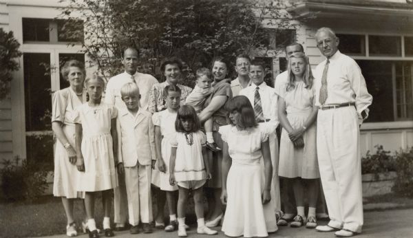 Outdoor group portrait of members of the Brumder family at the celebration of the forty-fifth wedding anniversary of George Frederick Brumder (1878-1961), far right, and Thekla Henrietta Wollaeger Brumder (1879-1950), far left. The well-dressed group is standing in front of the porch of G.F. Brumder's home on Pine Lake. See public note for identification of other family members and their relationship to George F. and Thekla Brumder. 