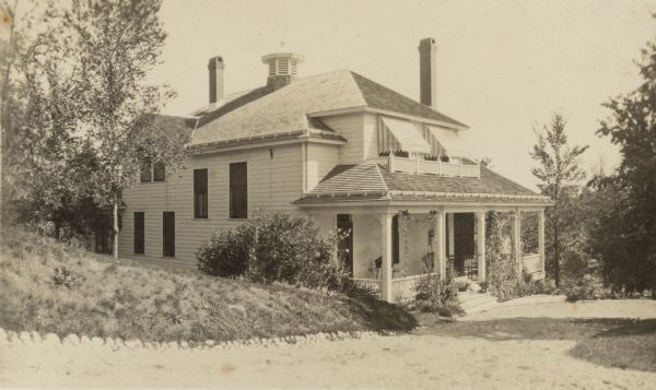 Three-quarter view from road of a wood frame, one and one half story house with a cupola and two tall chimneys. There is a full-length front porch on the main floor and large striped awnings on the front second-story windows on a balcony. Vines are growing on the porch and there is a picnic basket at the top of the steps. A glimpse of the lake is seen through the open porch. The house, the residence of George F. Brumder, was later enlarged and remodeled.