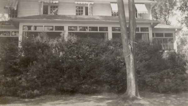 View of George F. Brumder's Pine Lake house, with a screened porch on the far right and enclosed porches left and center. Several of the windows on the upper level are open, with awnings above them. Shrubbery obscures the lower porch windows.