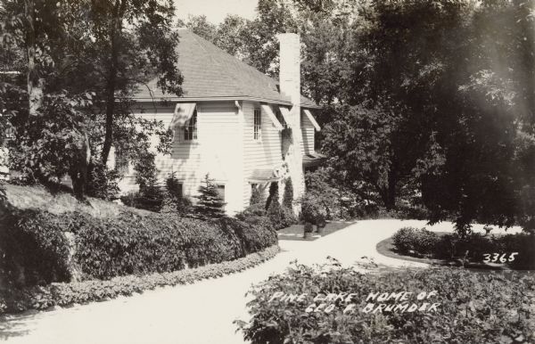 Photographic postcard view of a house from the driveway, which slopes down to the two-story wood frame building. Facing the driveway is an entryway which has a small roof over it, and a large chimney. The curved driveway is lined with small plants and ground cover. Caption reads: "Pine Lake Home of Geo F. Brumder." 