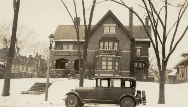 View across street towards an automobile parked in front of the George F. Brumder residence, which was built in 1910 on the corner of Grand (now Wisconsin) Avenue and 31st Street. The two and one-half story brick home, on a low hill rising above the sidewalk, features a large porch on the left and a prominent first story bay window, with a similar but smaller window on the second story. The house has three large chimneys, and a curved wall in the corner on the right. On the left, there are large houses facing 31st Street. A lamppost is standing near the sidewalk and steps leading up to the house; snow is covering the ground.