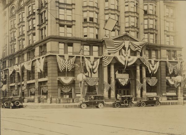 View from intersection towards a taxi and several other automobiles parked in front of the Brumder Building. Flags and flag-inspired bunting decorate the facades and lampposts. A sign over the main entrance reads: "Welcome A.A.C. of W. [Associated Advertising Clubs of the World] Milwaukee Herold and German National Farm Weeklies." Front windows to the left of the entry have signs identifying the National Bank of Commerce; those on the right have signs for the Milwaukee Herold and German National Farm Weeklies. A stairway on the left at the corner leads to a barbershop and bathhouse below street level.