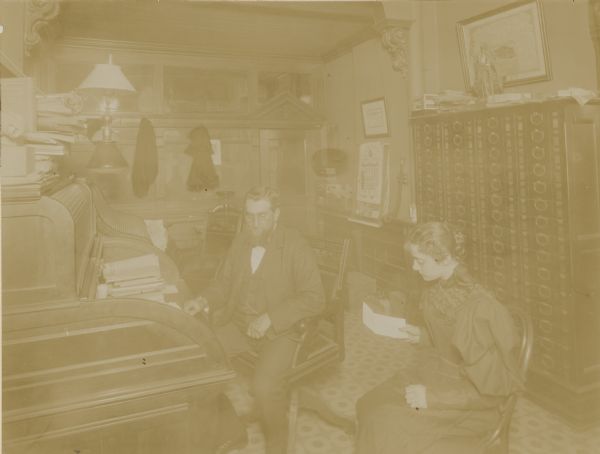 George Brumder, in a three-piece suit and wearing pince-nez eyeglasses, is sitting in a swivel chair at a roll top desk. His unidentified secretary is sitting near him in a bentwood chair, reading a note. There are file cabinets along the wall on the right, and a transitional lamp, fitted for both gas and electricity, is over the desk. A speaking tube is hanging on the wall behind Brumder, next to a large calendar. The far wall has interior windows and a door with detailed woodwork.  