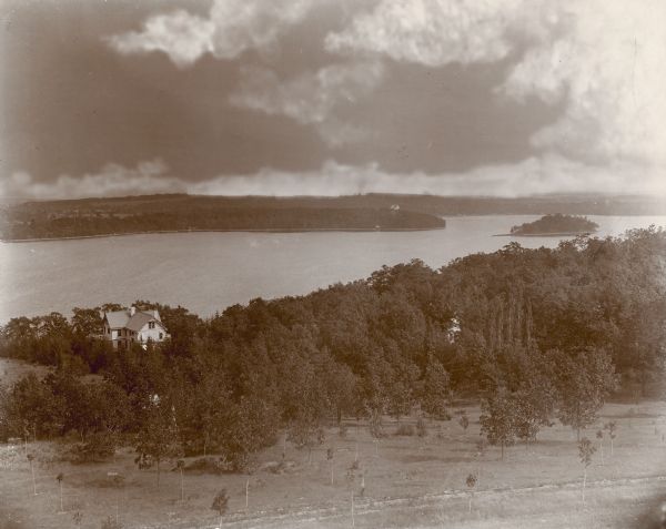 An elevated view of Pine Lake and the George Brumder summer house, taken from an observation tower built on a hill on the Brumder property. Many young trees have been planted in the foreground. The water storage tank for the house is visible through the trees near the house. Obscured by trees at the right is a glimpse of the caretaker's house.