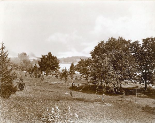 A view of the Pine Lake property of George Brumder, with the boathouse at far left. The smoke above the boathouse is from a steam water pump. There is a hose with sprinklers connected to it in the center of the lawn area. An unidentified man wearing a straw hat is standing with one foot on a wire bench conversing with Hugo Maerker, who is sitting in a canvas deck chair in the shade of trees. At far right is another bench, a hammock and two chairs. Pine Lake is in the background. On the reverse of the photograph is written: "View of lawn and boathouse with steam operated water pump. Taken from present walk leading from Emma Brumder's and Gertrude Merker Winkler's home. In distant trees to far left note Thekla P. Brumder's present home (1960)."