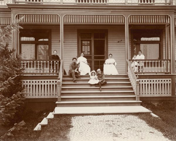 Members of George Brumder's family posing on the porch of the summer house on Pine Lake. They are, from left, Henriette Brandhorst (Mrs. George) Brumder; Hugo C. Maercker; unidentified; Gertrude Merker and Herbert Paul Brumder (the youngest son of George Brumder), both sitting on the steps; unidentified, possibly Amalie Christine Brumder; and Ida Brumder Merker, wife of Hugo Maercker, holding son Alfred Merker. Ornate fretwork and turned spindles adorn the porch. The steps lead to a stone walkway.
