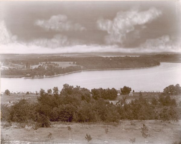 An elevated view, looking east, from the wooden tower on George Brumder's Pine Lake property, showing Pine Lake in the center and Beaver Lake beyond. There are young, newly planted trees in the foreground and, right of center, garden beds. An iron gazebo or garden house is standing between the gardens and the lake, and there is a small building with a tall smokestack on the shore. A note on the reverse of the photograph states: "Note George Brumder's garden, iron garden house and boathouse. Also the Friend house and windmill across the lake."