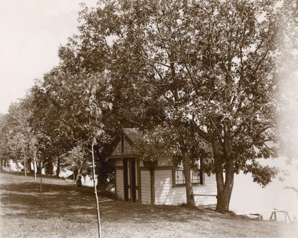 A small bathhouse is standing among trees on the shore of Pine Lake on George Brumder's property. There is ornate fretwork in the gable ends. A sign on the front reads: "Bathhouse"; "Ladies" is written on another sign on the door. Brumder's son-in-law, Hugo C. Maercker, is seen in profile to the left of the structure. He appears to be sitting at the slope of the shoreline. On the right next to the bathhouse is a plank nailed between two trees to form a bench. A simple pier or diving platform is over the water just beyond the bench to the right. 