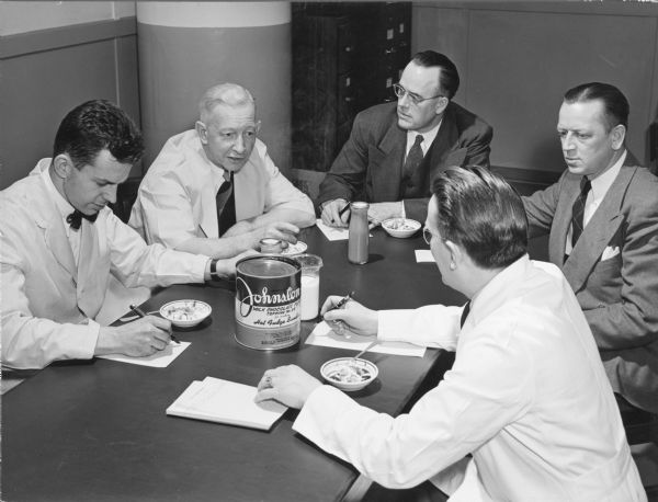 Five men, each with a small dish in front of them, are sitting around a table in the chemistry laboratory of the Robert A. Johnston Company. Several of the men appear to be making notes, and three of the men are wearing white lab coats. An open tin of Johnston Milk Chocolate Fudge Topping is sitting at the center of the table. There is also a beaker of milk and two unopened chocolate milk bottles on the table. A hand written note on the reverse of the photograph identifies the "RAJ Co. Chemistry Lab," and the men, clockwise from left, as: "Ned, N.J. Russel, Petran, Bill Manschot, and Dan Wing." Ned is Edward Crandall Johnston (1912-1955), the grandson of the company's founder Robert A. Johnston, and the son of Walter Vail Johnston. Robert A. Johnston Co. made cookies, candy, chocolate and other foods in Milwaukee, and later in West Allis, from 1899 until 1969.
