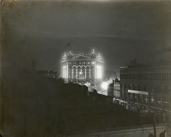 Elevated night view of the eight story Germania Building, which is illuminated with a multitude of individual electric light bulbs outlining the windows and other architectural details. The trapezoidal building has four domes at the corners. A statue of "Germania" stands on a pediment at the fourth floor level and lifts an illuminated torch in her right hand. A small triangular building facing the front of the Germania Building is named, appropriately, The Triangle, and advertises the Cream City Brewing Co. on large signs at roof level. There are brick buildings on the right with signs for "Leather and Findings," "Western Bottlers Supply Co.," and "Beckerman & Mayer Mfgrs of Cloth Hats and Caps."
