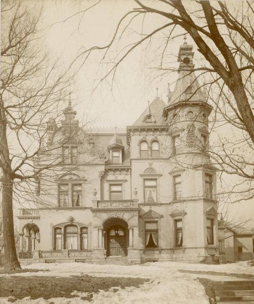View across lawn, covered partially with snow, towards the Grand Avenue (later Wisconsin Avenue) facade of the George Brumder home at the corner of 18th Street. The three-story brick Queen Anne Style home features several porches, a turret, plaster ornamentation and Flemish Style details. There is a decorative iron fence on the roof ridge. Draperies are in the windows.  