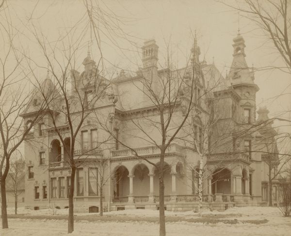 View across lawn, covered partially with snow, towards the 18th Street side of the George Brumder mansion on Grand (later Wisconsin) Avenue. The three-story brick Queen Anne Style home has multiple porches, a turret, plaster ornamentation and Flemish Style gables. A chimney rises high above the roof. Two other Grand Avenue mansions are glimpsed at far right.