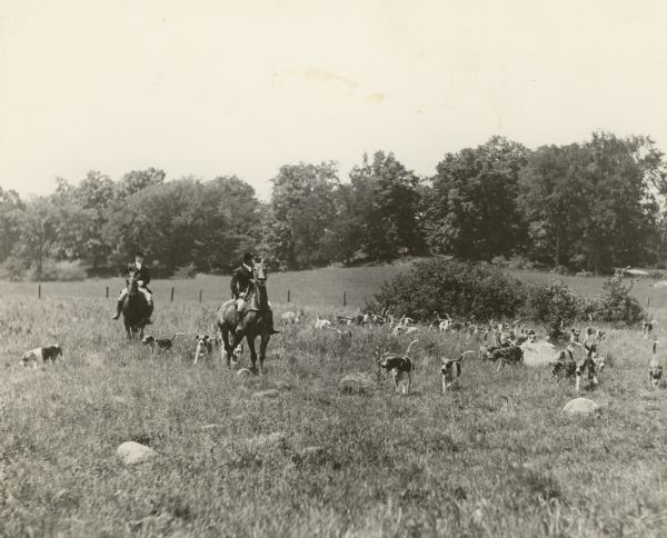 Two unidentified men wearing traditional hunting dress, including hunt caps, are riding horses with a pack of foxhounds in a rocky meadow. There is a fence and trees in the background. A handwritten caption identifies the Lord Fitzwilliam Pack of Washington County. The caption also includes: "John Cudahy M.F.H. August 1931."   
