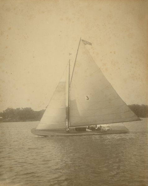 The Class A, shovel-nosed sailboat, the <i>Aderyn</i> sailing on Lac La Belle. A handwritten caption on the reverse of the photograph identifies Herman Otto Brumder, skipper; his brother George F. Brumder, sheet; Adolph Rietbrock, prone on starboar deck; and Bob Manegold, at mast. The <i>Aderyn</i> was built by Jones and LaBorde boatbuilders in Oshkosh, and owned by George Brumder, Milwaukee publisher and businessman whose summer home was on Pine Lake. The <i>Aderyn</i> won the Inland Lake Yachting Association Regatta on Pewaukee Lake in August 1902.