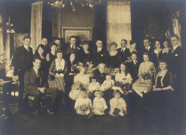 Henriette Brandhorst Brumder, sitting in the center holding two of her infant grandchildren, posing with family members for a portrait on her 80th birthday. The family has gathered in the living room of the Brumder house at the corner of Grand (now Wisconsin) Avenue and 18th Street. Several of the children are holding bouquets. The furnishings reflect the time period in which the house was built, ca. 1895. There are heavy drapes with lace sheers on the windows, and transitional chandeliers, fitted for both gas and electric lights. Henriette was the widow of Milwaukee publisher and businessman George Brumder.  