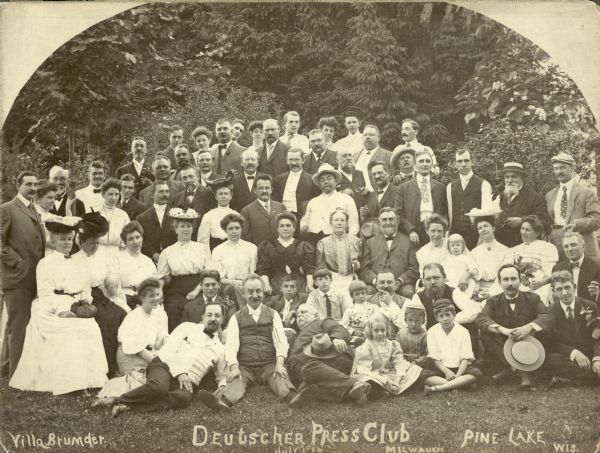 An informal group portrait of members of the Milwaukee Deutscher Press Club and others, taken at Villa Henriette (mistakenly labeled "Villa Brumder"), the Pine Lake summer home of publisher George Brumder.  Brumder, with beard and bow tie, is seated next to his wife Henriette, who is holding an umbrella, just right of center. Their daughter Amalie, holding her daughter Dorothea (Dodi), is to the right of George. George's son William C. Brumder is standing at extreme left; behind him is his sister Ida Brumder Merker. Another of George's sons, Herman O. Brumder, is in the back row, right, wearing a short billed German style student cap. Gustave Haas, managing editor of Brumder's newspaper, The Germania, is in front and to the right of Herman.  Emil Von Steinitz, another editor of The Germania, is in the center, third row from the rear, wearing pince nez glasses on a string, and no necktie. The wide-eyed gentleman sitting up in the front row is identified as "Koenig," possibly Charles H. Koenig, president of the Mount Goram Mining and Milling Company.