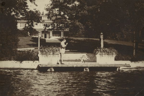 Herman Otto Brumder (1880-1950) standing on the boat landing of the Pine Lake home built by his father, George Brumder.  Herman is holding a treat for his dog Muggsy. There are large planters on the landing, and two lights on poles. The large two-story wood frame house has an attic dormer and a one-story porch facing the lake.  