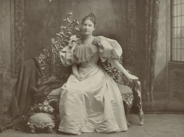 Full-length studio portrait in front of a painted backdrop of Emma Brumder (1870-1962), daughter of George and Henriette Brandhorst Brumder. She is sitting on a throne-like love seat which has a matching footstool, and is wearing a full-length dress with balloon sleeves and long white gloves.