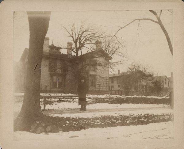 A view of the south (Wells Street) side of the two-story brick Italianate house at the corner of 10th Street purchased by George Brumder in 1882. An unidentified man, possibly William C. Brumder, is wearing an overcoat and hat and posing on the sidewalk along Wells Street. The house has three large chimneys and a Belvedere with a spire. There is a first floor bay window with decorative ironwork, and a trellis on the wall at far right. The building to the right of the house is a barn.
