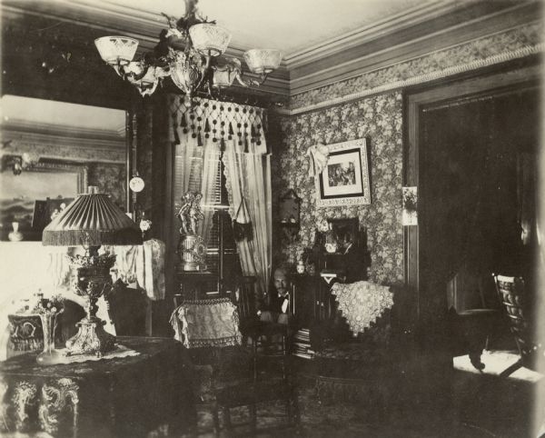 An interior view of the music room of the George Brumder home at the corner of Wells and 10th Streets. The room has a high ceiling and is decorated in Victorian style with elaborate wall coverings and draperies, and a large gas-lit chandelier. William C. Brumder, partially obscured by a chair, is crouching in the corner of the room.