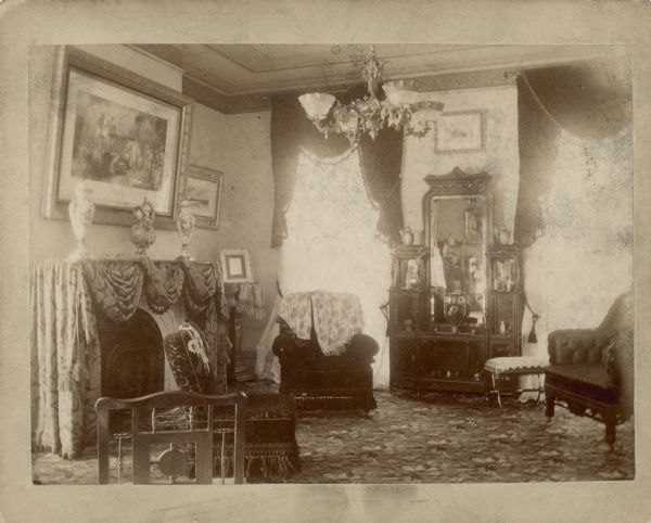 A view of the southwest corner of the living room of the George Brumder house at the corner of 10th and Wells Streets. An ornate Victorian etagere stands between two windows against the far wall. On the left is a fireplace with a cloth draped over the mantle, where two urns and a ewer are displayed. There are framed paintings or prints on the walls and a gas-lit chandelier overhead.