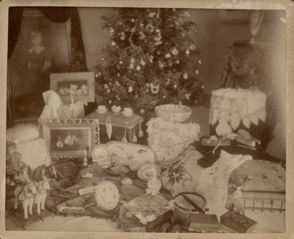 An assortment of Christmas gifts are arranged around the base of a Christmas tree in the living room of the George Brumder house at the corner of 10th and Wells Street. A corner of the draped fireplace mantle is on the far right. Gifts include a doll, toy horse-drawn wagon, and drum. Also on display are books, a dresser set, several embroidered pillows, a large fish platter and several framed prints.