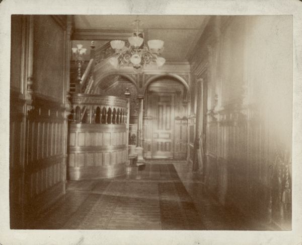A view of the wood paneled entry hall of the George Brumder residence at the corner of 18th Street and Grand (later Wisconsin) Avenue. There are carpets on the floor and an overhead chandelier fitted for both gas and electricity. There is a gas light fixture on a newel post of the stairs.