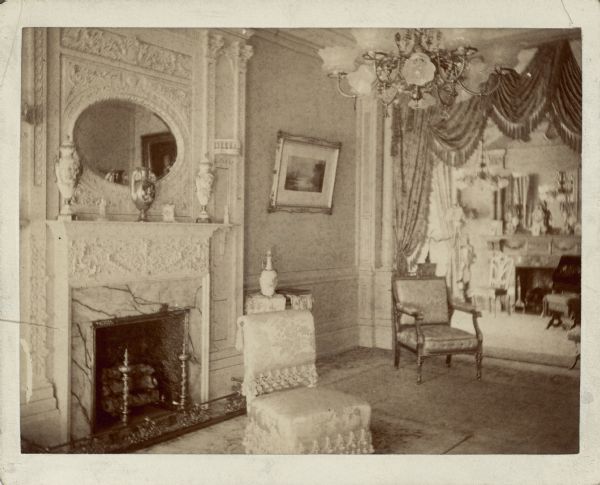 A view of the parlor (foreground) and music room of the George Brumder residence at the corner of Grand (later Wisconsin) Avenue and 18th Street. Both rooms feature a fireplace and chandelier fitted for both gas and electricity. There are three urns on the mantle in the parlor, and a framed print or painting on the wall. Ornate draperies frame the wide doorway between the two rooms.