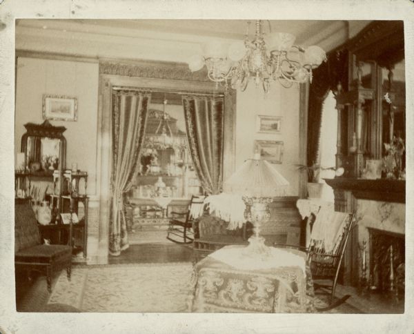 The living room of the George Brumder residence at the corner of Grand (later Wisconsin) Avenue and 18th Street. There is a fireplace on the right. An ornate etagere stands at left, next to a doorway into the library. There are drapes in the doorway. Overhead is a chandelier fitted for both gas and electric lights. There is a table lamp in the foreground.  