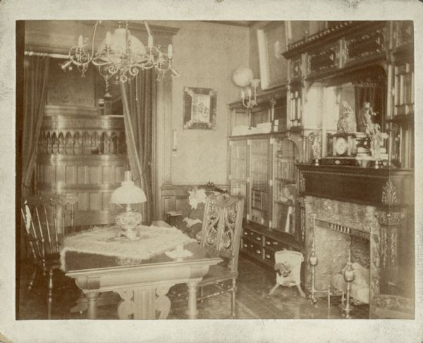 A view of the library in the George Brumder residence at the corner of Grand (later Wisconsin) Avenue and 18th Street. There is a fireplace with mantel clock on the right, and a library table with two chairs in the foreground. A built in bookcase with lower drawers is to the left of the fireplace. An overhead chandelier and wall sconce are fitted for both gas and electric lights. A doorway with drapes affords a view of the main hall with its wood paneling.