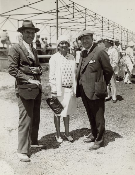 Walter Vail Johnston, left, posing with an unidentified woman and man near the grandstand at the Milwaukee Hunt Club Horse Show. The men are wearing suits and ties; the woman is wearing an outfit with a polka dot top. Johnston and the woman are both wearing spectator shoes.