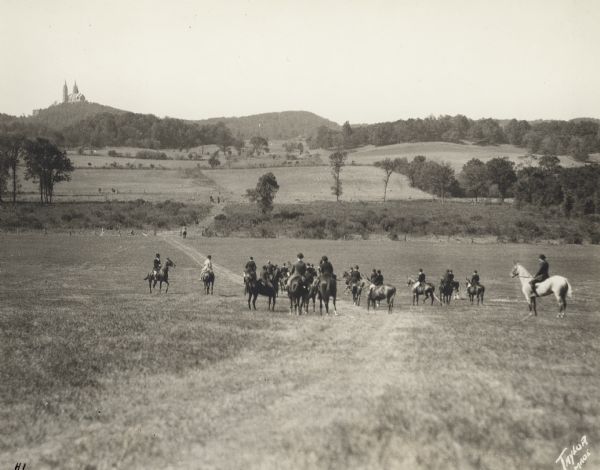 A group of men and women on horseback pausing during a fox hunt. They are in traditional fox hunting attire. The Basilica of Holy Hill is in the distance. On the reverse of the photograph is written: "Holy Hill Break-Off Group."