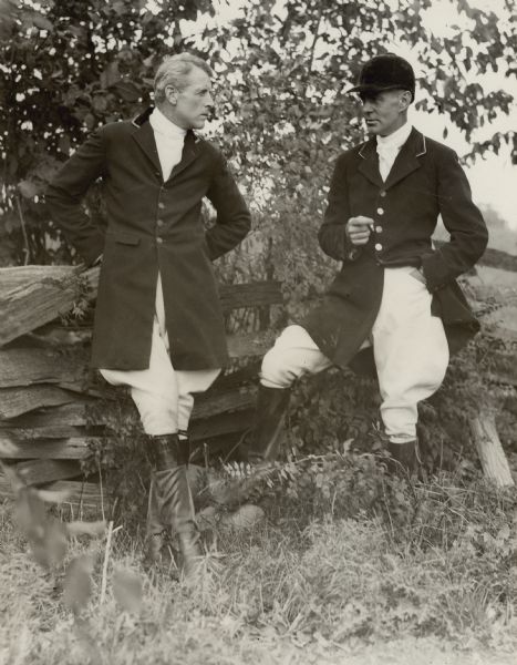 John Clarence Cudahy, left, and Walter Vail Johnston are resting against a split rail fence during the Washington County Fox Hunt. Both were members of the Milwaukee Hunt Club. The men are wearing traditional fox hunting attire; Johnston is wearing his cap and is smoking a cigarette.