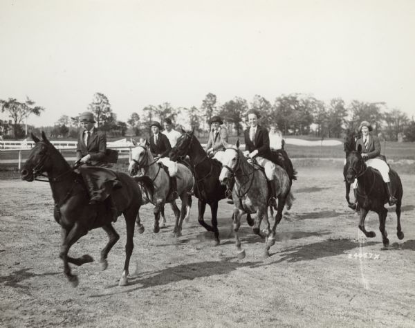 June Ellen Johnston, far left, leading a group of young people on horseback at the Milwaukee Hunt Club. She is wearing a hat, jacket and necktie, and a long skirt as she is riding sidesaddle. The other young women in the group are wearing jodhpurs and are riding astride the horses. There is a golf course in the background.
